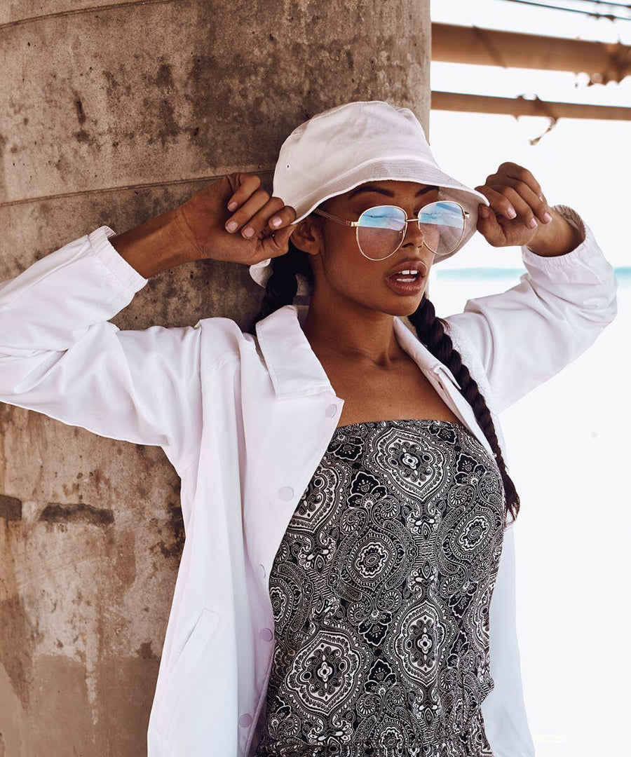 A woman stands against a concrete wall, adjusting her Flexfit By Yupoong YP039. She is wearing mirrored sunglasses, a white jacket, and a patterned dress.