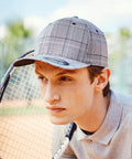 A person wearing a checkered cap, specifically the YP041 by Flexfit By Yupoong, and a gray polo shirt holds a tennis racket, looking intently to the side, set against an outdoor tennis court background.