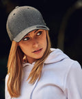 A person with long hair wearing a gray Flexfit By Yupoong YP044 baseball cap and a white hoodie looks to the side against a dark background.