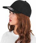 A woman with long, wavy brown hair wears a black Flexfit By Yupoong YP054 baseball cap and a white top, looking to the left.