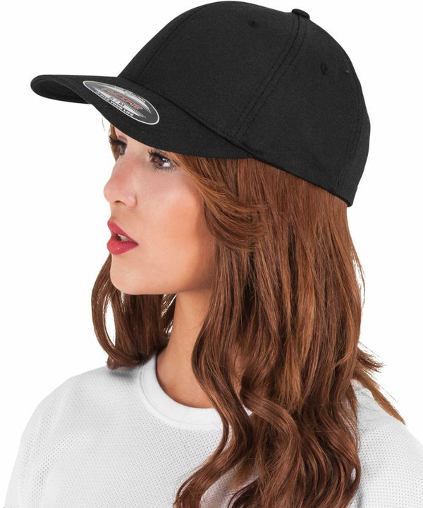 A woman with long, wavy brown hair wears a black Flexfit By Yupoong YP054 baseball cap and a white top, looking to the left.