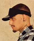 Side profile of a person in a Flexfit By Yupoong YP059 and plaid shirt, with visible ear piercings, neck tattoos, and a teardrop tattoo near the left eye. The background is a beige textured wall.
