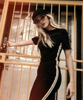 Person in a Flexfit By Yupoong YP064 black cap and black dress with colorful side stripes, leaning against a yellow metal gate, looking at the camera.
