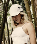 Person wearing a white cap (Flexfit By Yupoong YP065) and a white sleeveless top, standing in a forested area with bamboo in the background.