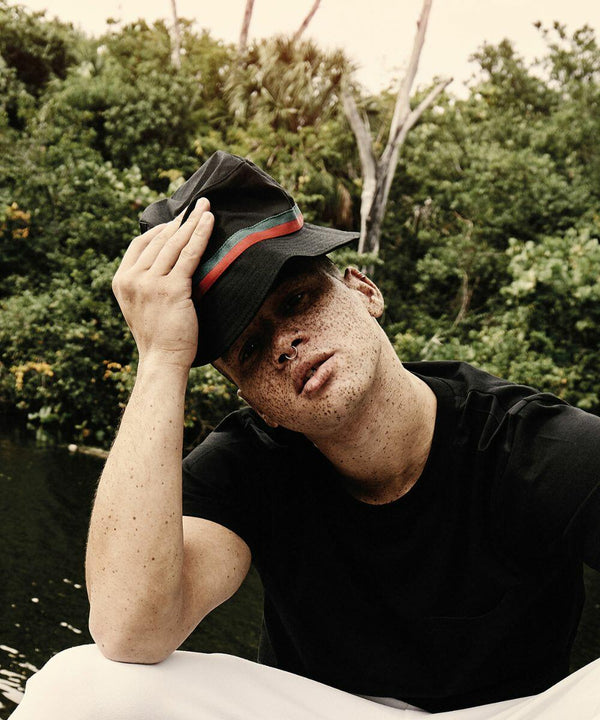 Person wearing a dark Flexfit By Yupoong YP072 with a green and red band, black shirt, and white pants, sitting outdoors near a body of water with a backdrop of lush greenery.