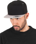 A bearded man in a black crew-neck shirt and a Flexfit by Yupoong Classic 5-panel snapback (6007T) with a grey brim looks slightly left against a plain white background.