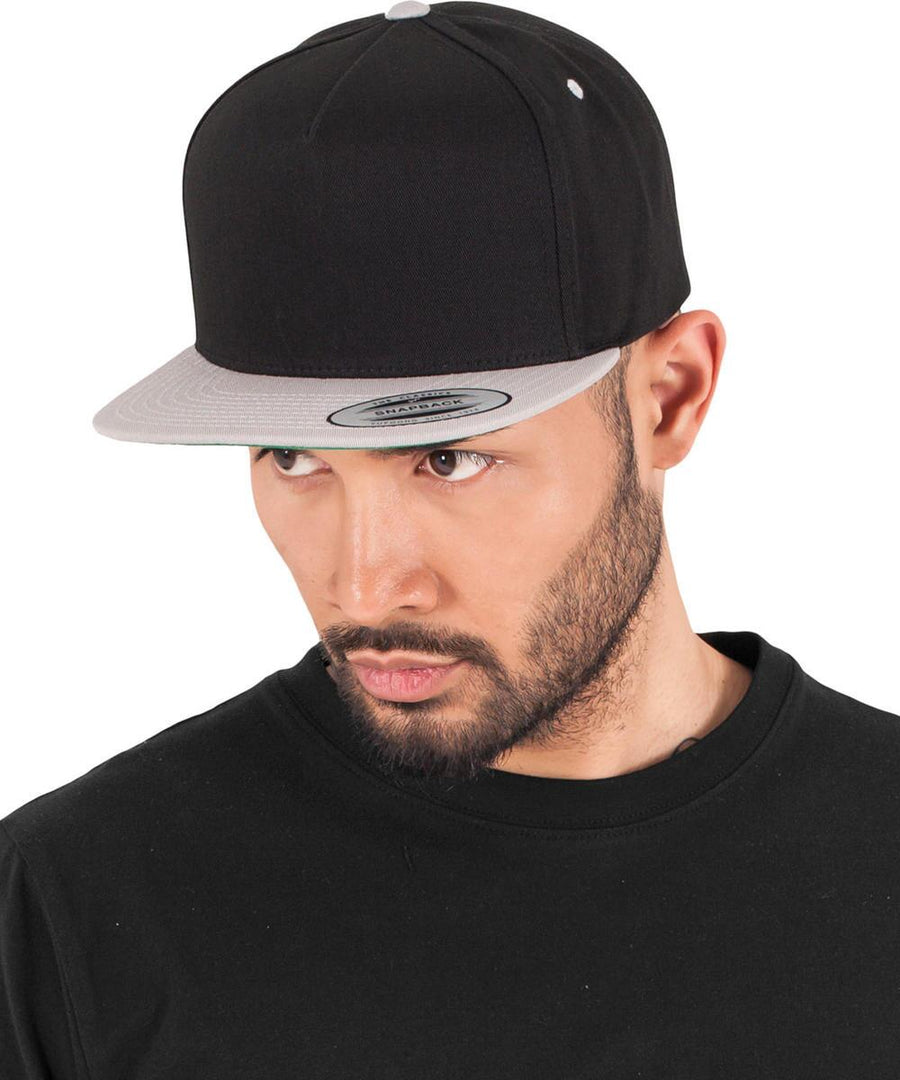 A bearded man in a black crew-neck shirt and a Flexfit by Yupoong Classic 5-panel snapback (6007T) with a grey brim looks slightly left against a plain white background.