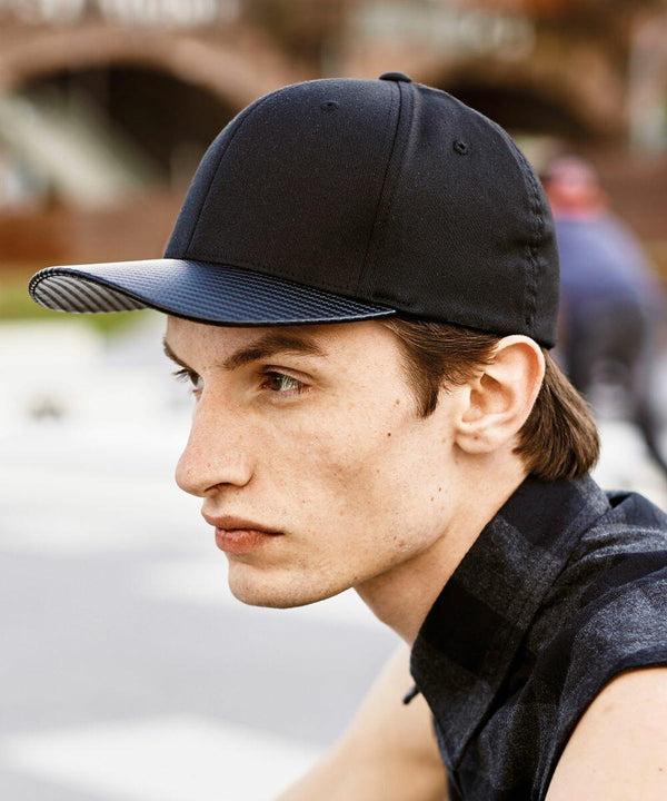 A man with light skin and brown hair is seen in a profile view wearing a black and blue Flexfit By Yupoong YP079 cap. He is outdoors, sitting on a bench, with blurred buildings and people in the background.