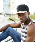 A young man wearing a black Flexfit By Yupoong YP082 and striped tank top sits outdoors with a serious expression, holding onto a bicycle.