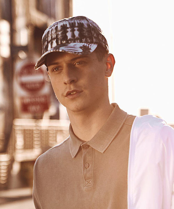 A young man wearing a Flexfit By Yupoong YP099 and a polo shirt stands outdoors with blurred street signs in the background.