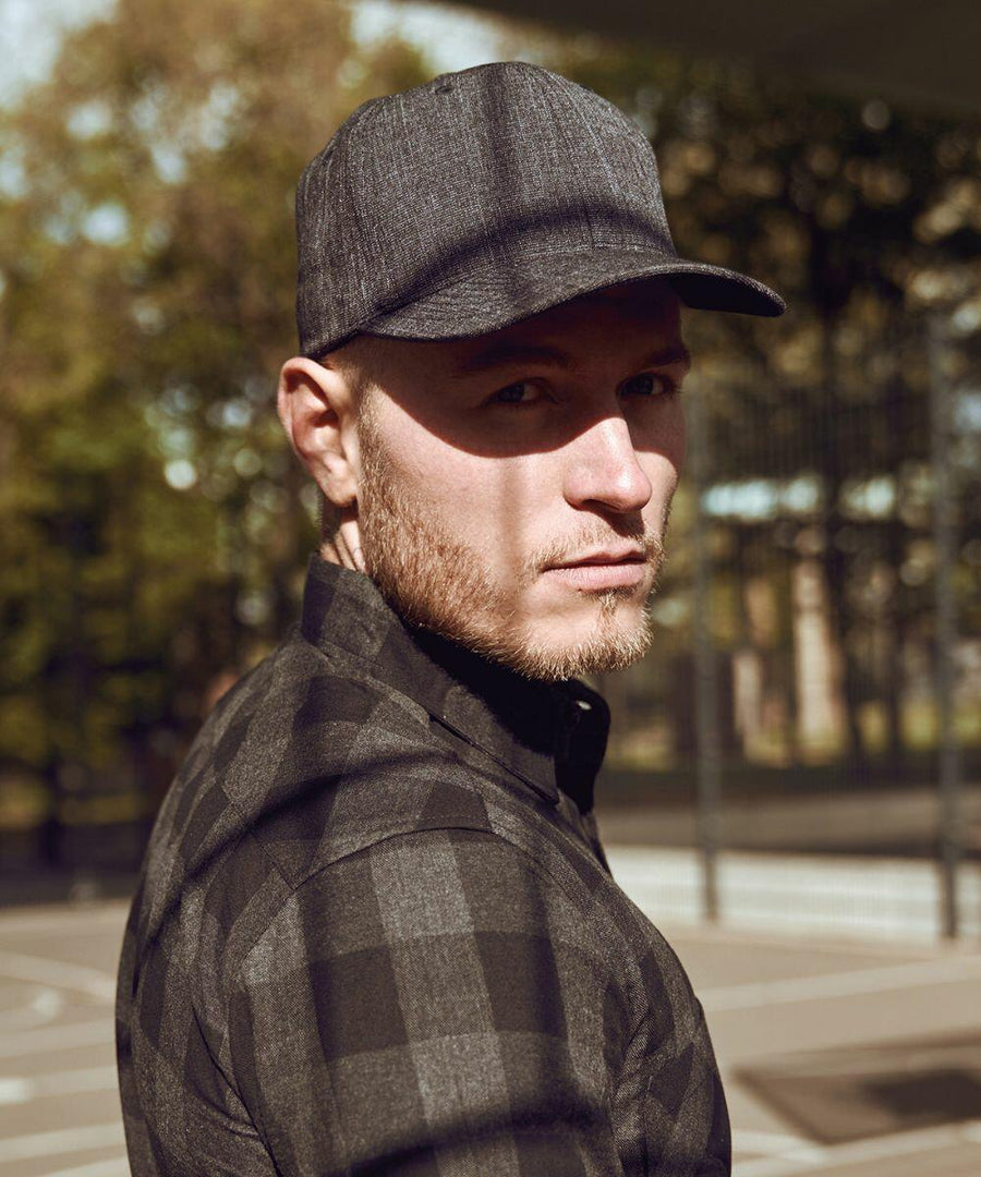 Man wearing a dark plaid shirt and gray cap, specifically the YP105 by Flexfit By Yupoong, looking over his shoulder, standing outdoors in a sunlit area with trees and fencing in the background.
