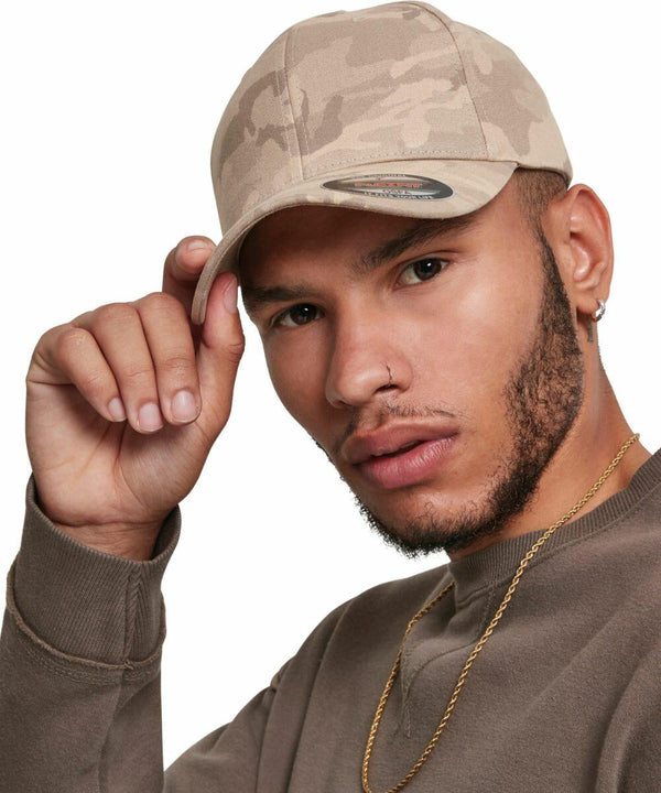 A person wearing a beige camouflage Flexfit By Yupoong YP110, a nose ring, and a gold chain, looking at the camera while adjusting the cap with one hand.
