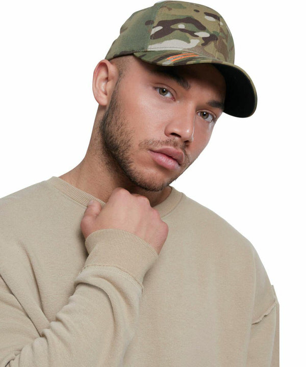 A man wearing a beige sweatshirt and a Flexfit By Yupoong YP112 camouflage baseball cap poses with his hand on his neck against a white background.