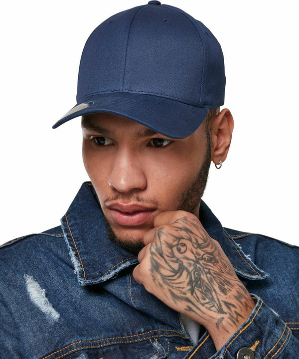 A person wearing a navy blue YP113 baseball cap from Flexfit By Yupoong and a denim jacket, with a tattoo on their right hand, holds their chin with a thoughtful expression.