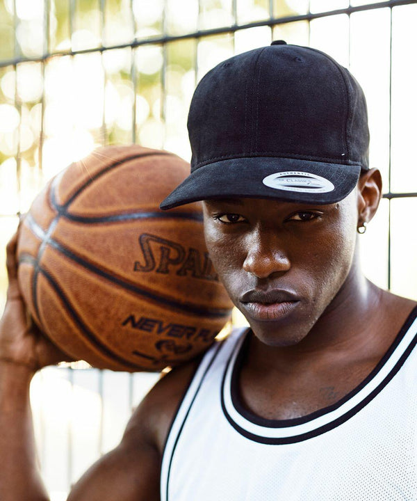 A person wearing a black Flexfit By Yupoong YP123 cap and white tank top holds a basketball on their shoulder while standing in front of a fence.