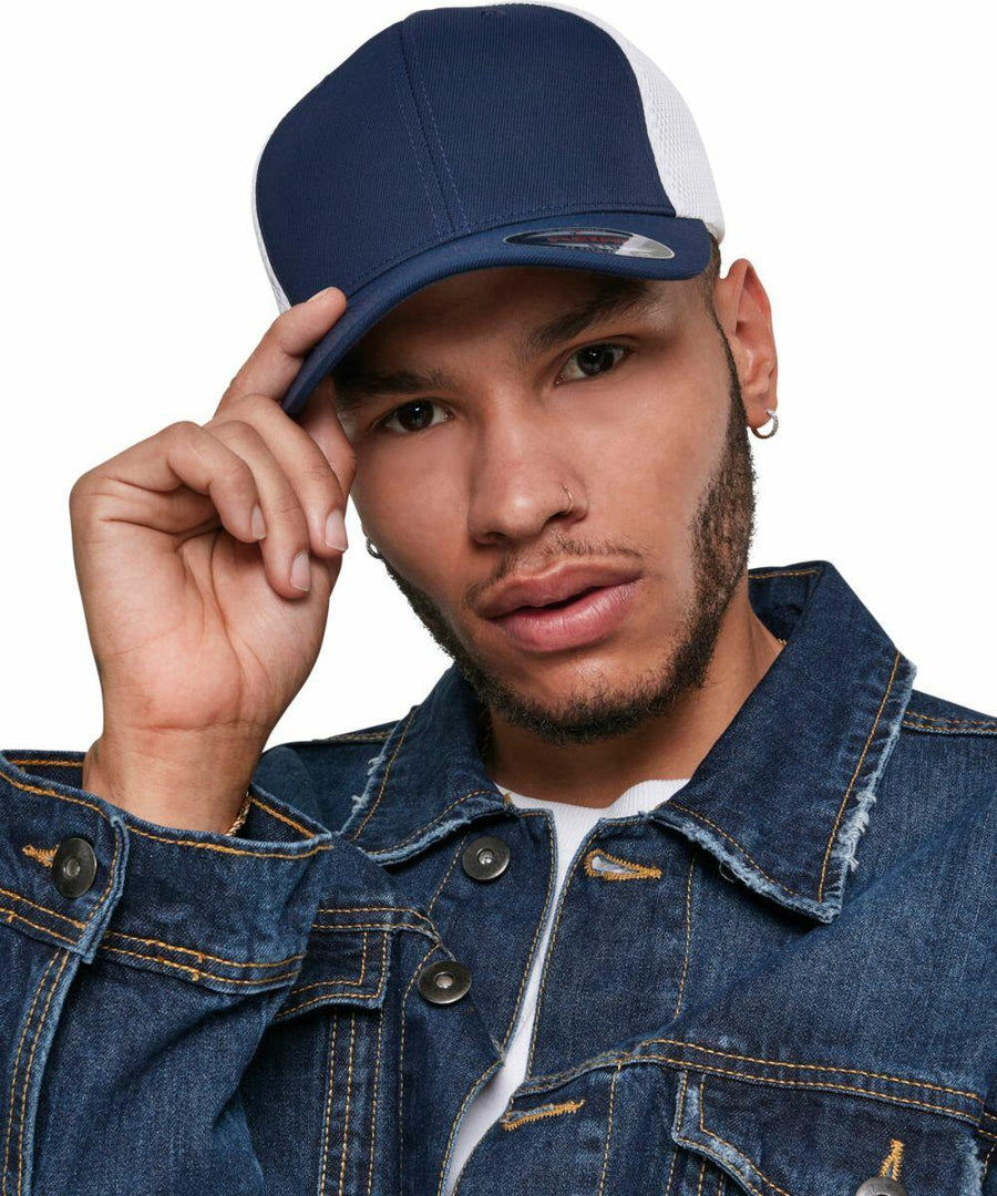 A person wearing a dark blue and white Flexfit By Yupoong YP128 cap, denim jacket, and white t-shirt looks at the camera while holding the brim of the cap with their right hand.