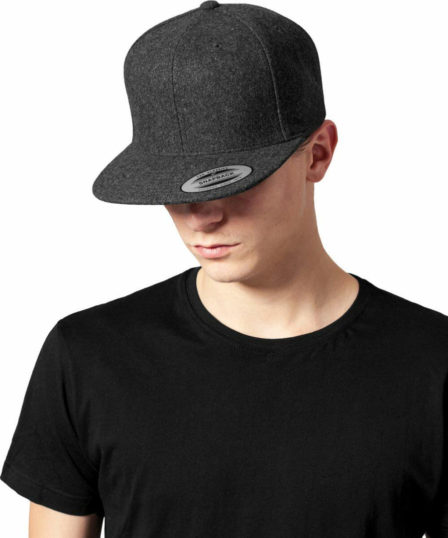 A person wearing a plain black t-shirt and a dark grey Flexfit By Yupoong YP132 flat-brimmed cap, looking downward, with the face partially obscured by the cap's brim.