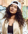 A person with long, curly hair wearing a white and beige jacket and a YP211 by Flexfit By Yupoong worn backwards, standing outdoors with a graffitied wall in the background.