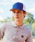 A young man outdoors wears a Flexfit by Yupoong Flexfit trucker mesh 2-tone (6511T) cap in blue and white, along with a light grey polo shirt. He looks slightly to the side with a neutral expression; trees and sky are blurred in the background.