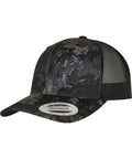The Flexfit by Yupoong YP Classics Veil Camo retro trucker cap (6606VC) features a black and grey camo pattern, curved brim, black mesh back panels, and a round manufacturer sticker on the brim.